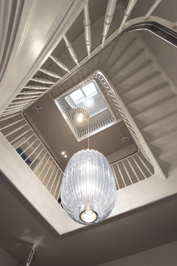 Blick von unten in ein hohes klassisches Treppenhaus. Im Treppenauge hängen drei große Leuchter auf unterschiedlichen Höhen